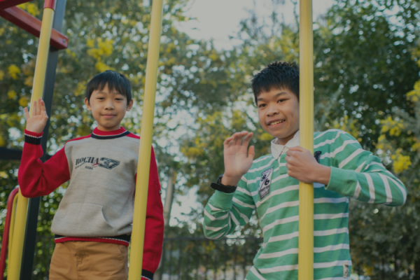 Two boys on play equipment waving at the camera. There are trees in the background.