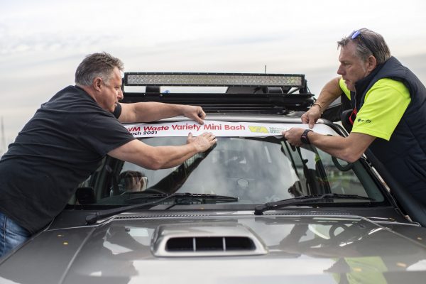 Two men leaning across a vehicle putting a sticker on the front windscreen