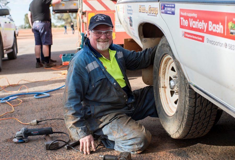 Man sitting on the ground wearing high-vis and working on the back wheel of a car that is mounted. 