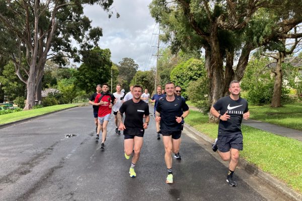 A group of people are running on a road, surrounded by greenery and foilage. The three at the front of the back are all dressed in black and smiling at the camera.
