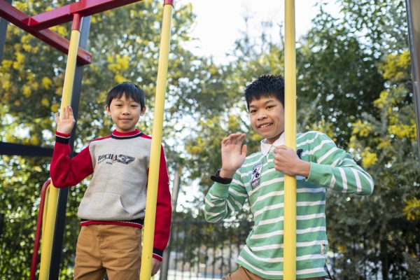 Two boys on play equipment waving at the camera. There are trees in the background.