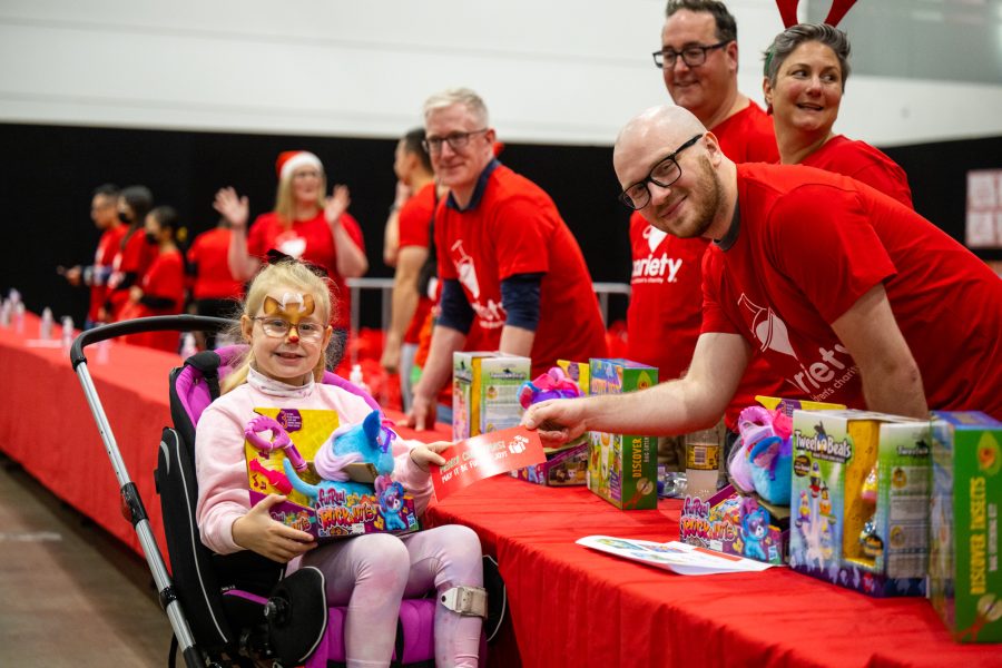 Young girl in wheelchair wearing reindeer face-paint receives a Christmas gift from a Variety volunteer